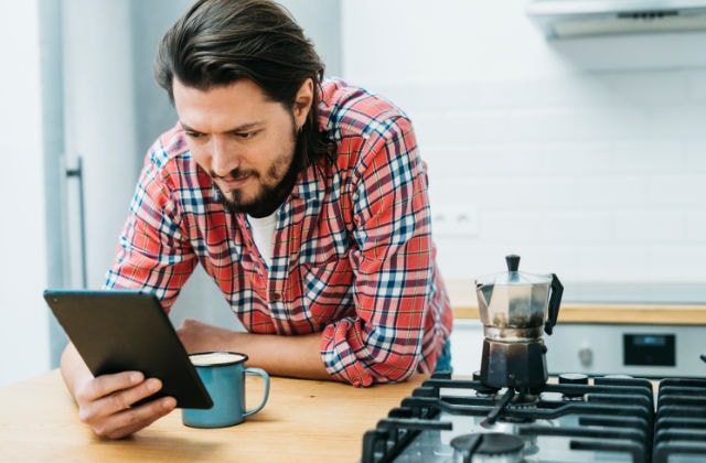Hombre leyendo sobre el tope del gas en una tablet mientas toma un café en la cocina